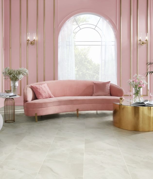 Living area with pink wainscoting, arched window, gold sconces, pink velvet sofa, and large gold coffee table topped with rose bouquet and wine glasses, with opalescent stone-look floor