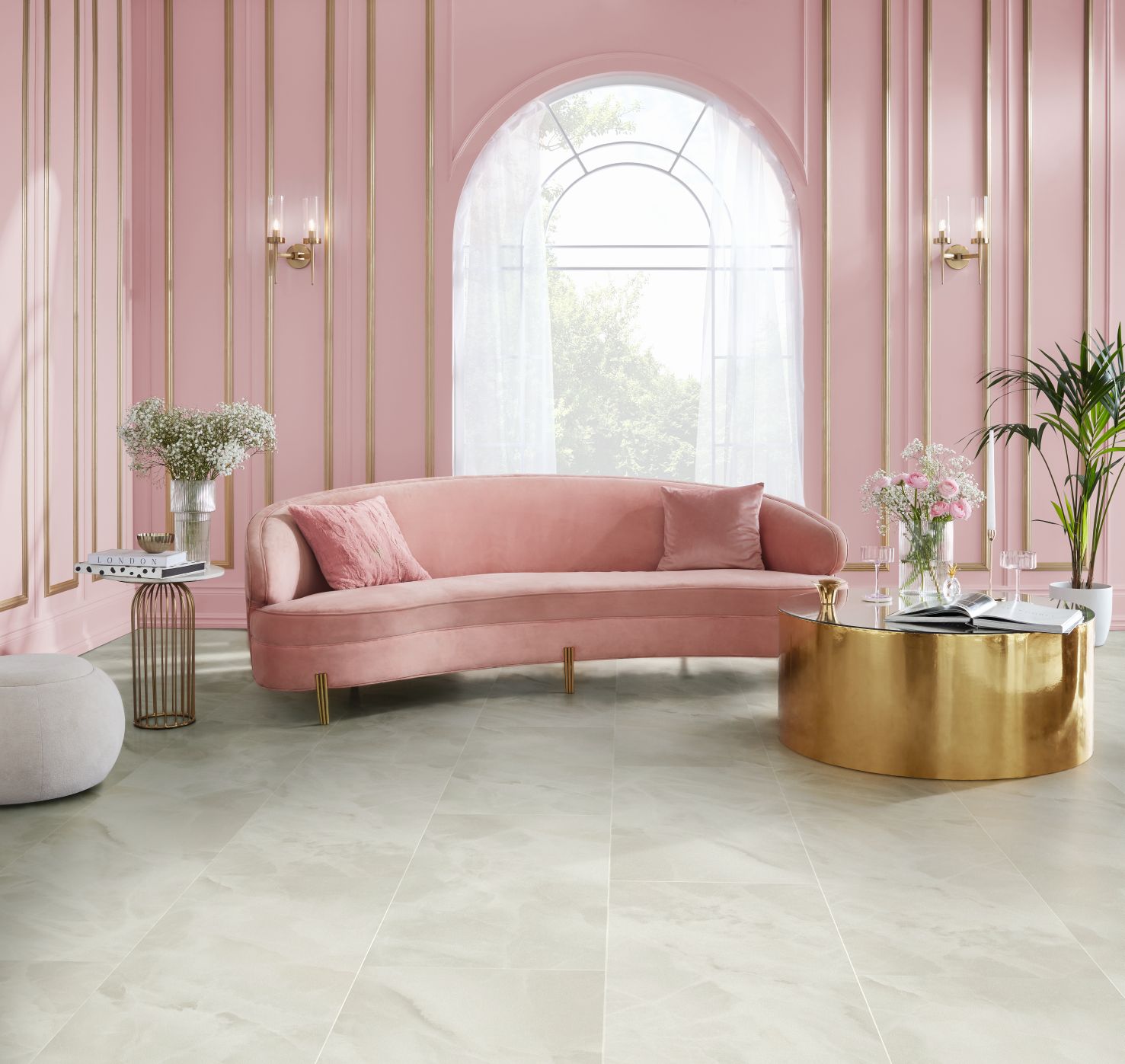 Living area with pink wainscoting, arched window, gold sconces, pink velvet sofa, and large gold coffee table topped with rose bouquet and wine glasses, with opalescent stone-look floor