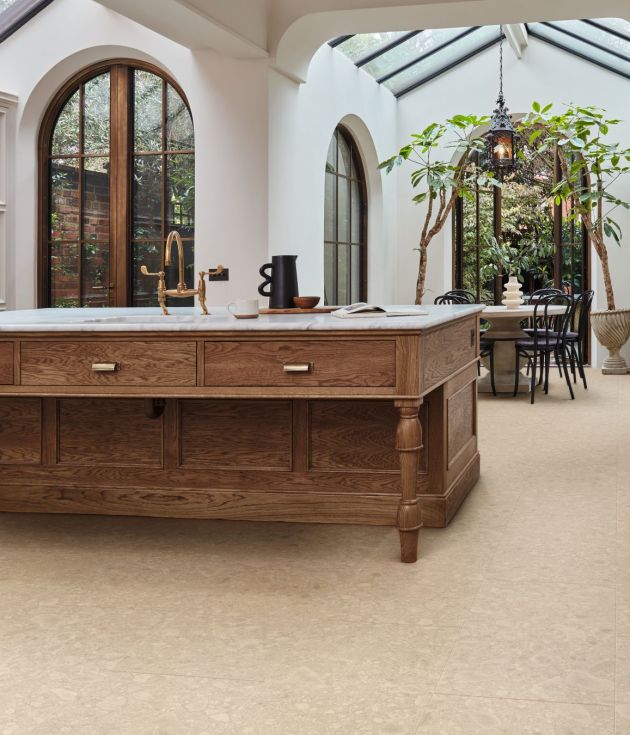 French country kitchen area, with exposed beams, arched doorframe, and large wooden island, with indoor trees flanking dining area in background, ecru wainscoting and taupe terrazzo floors