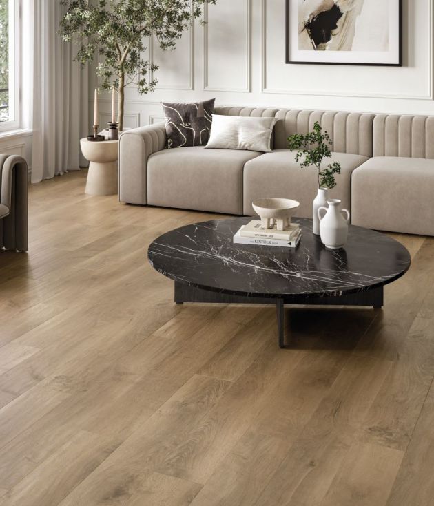 Calm neutral living space with modern couch, armchairs, black marble coffee table topped with pottery, with artwork and large houseplant on muted brown woodgrain floor