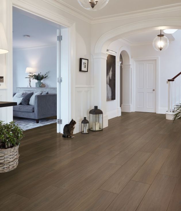 Refined and grounded hallway showing peek of seating area and staircase in background, with side table topped with dotted lamp, with potted plants, cat, and decorative candle lamps on chocolate-brown woodgrain floor