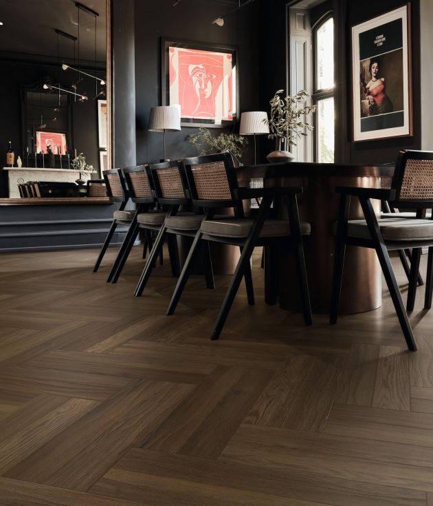 Dark and moody dining area with brown herringbone woodgrain floor, table and chairs, artwork, and peek of steps ascending to bar area in background