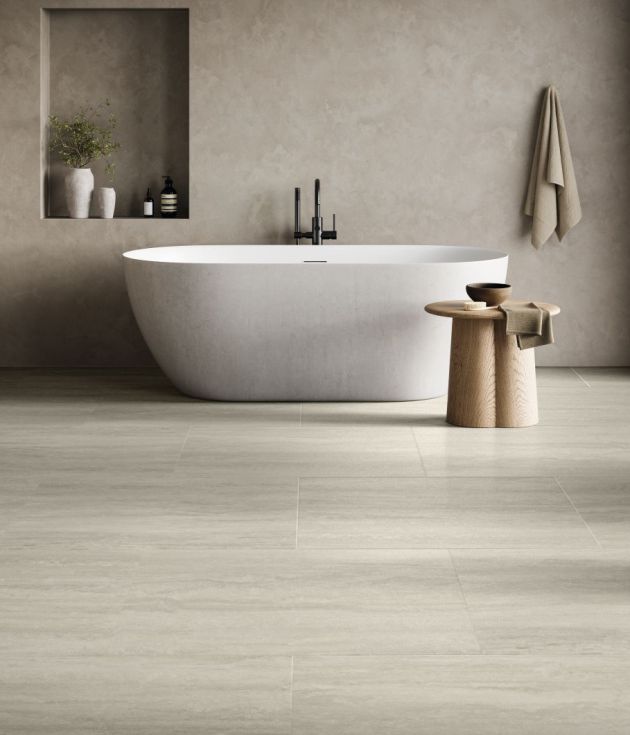 Bathroom featuring gray concrete-look wall with niche, hanging towel, and side table beside soaker bathtub, in room with gray travertine-style tile floor