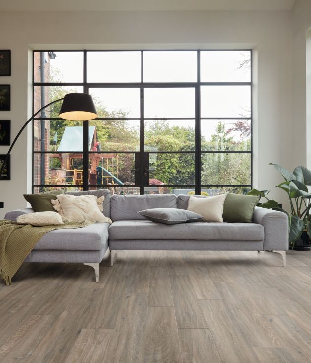 Living space with grayish-brown woodgrain floors, gray couch, arched floor lamp, and potted plant set before large windows with playground outside