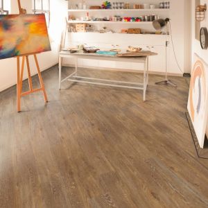Home office art space with painting on easel with table and floor lamp near built-in shelves packed with art supplies, in room with lime-washed brown woodgrain floor installed at a diagonal with a dark decorative design border
